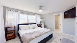 A bedroom with a bed, a nightstand, a lamp, a window with curtains, and a TV mounted on the wall.