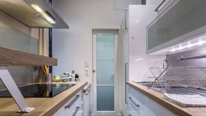 Une cuisine étroite avec un plan de travail noir, des placards blancs et une porte en verre donnant sur une autre pièce.