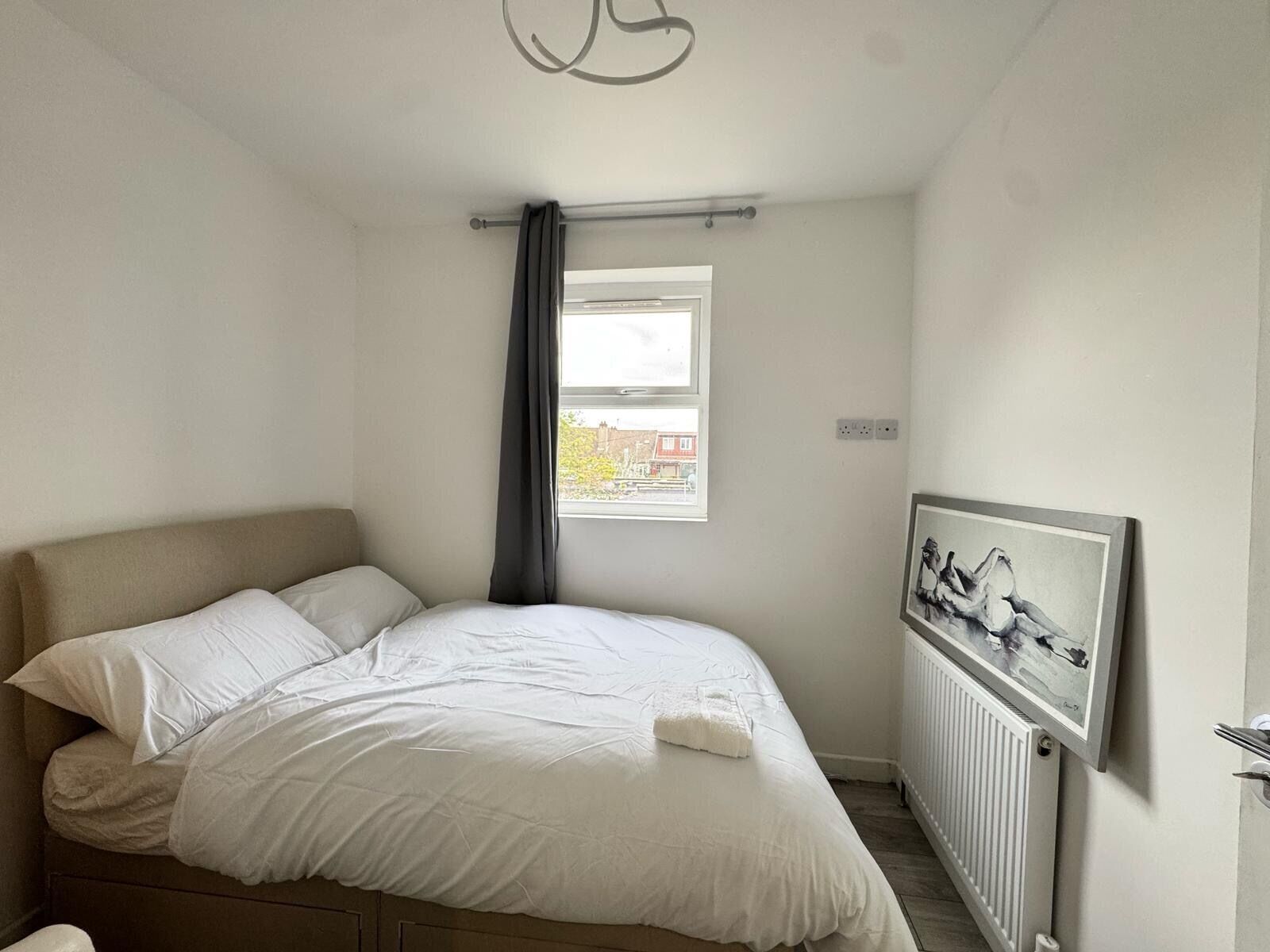 A bedroom with a bed, a window with a view of buildings, a radiator, and a wall-mounted artwork.