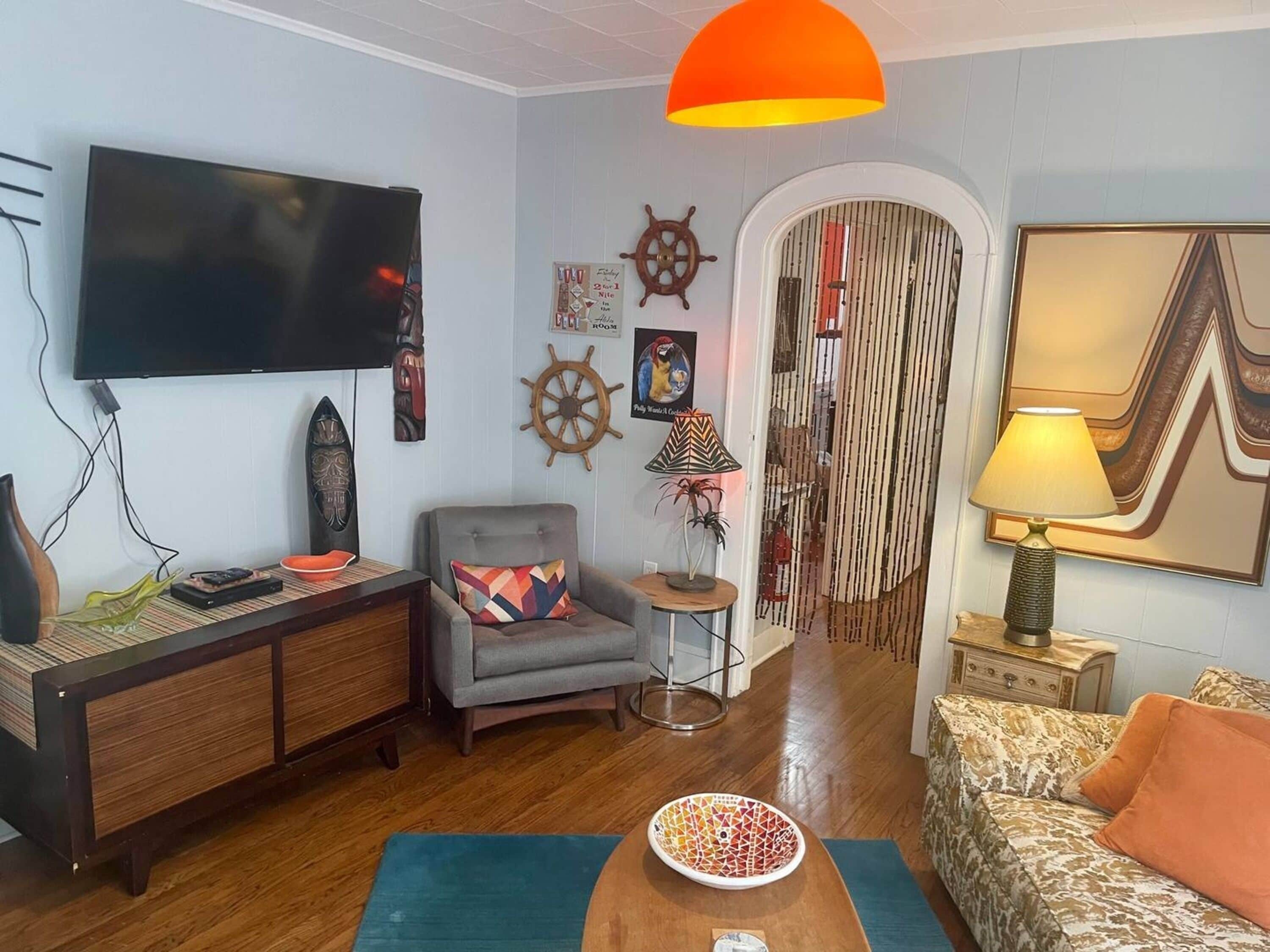 Un salon doté d’un téléviseur à écran plat, d’un fauteuil gris, d’un canapé à motifs, d’une table basse avec un saladier, et d’un grand luminaire suspendu orange.