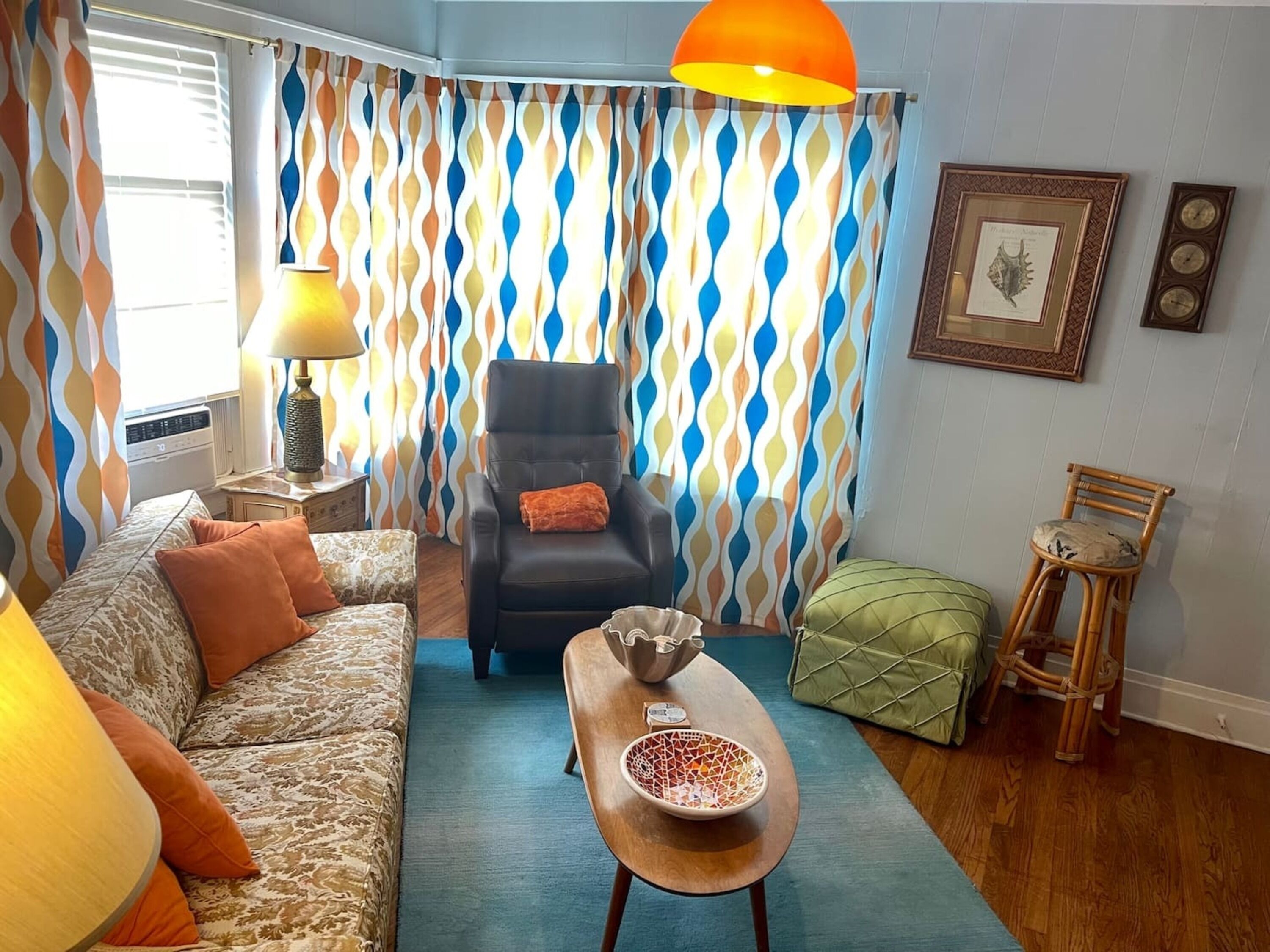 A living room with a patterned curtain, a sofa with cushions, a chair, a small table with a bowl, and a lamp.