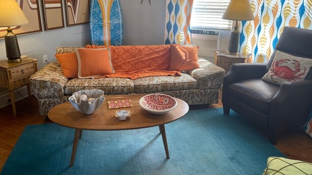 A living room with a patterned sofa, a coffee table, and a chair. There are surfboards on the wall, a lamp on a side table, and a window with curtains. - 1BR Coastal Escape (Galveston)