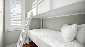 Uma cama beliche com roupa de cama branca e uma escada, uma janela com persianas e piso de madeira.