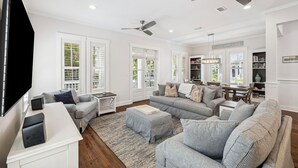 A spacious living room with a large flat-screen TV, grey sofas, a coffee table, and a bookshelf.