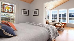 A bedroom with a large bed, two framed pictures, a wooden ceiling, and a view of a lake through large windows.