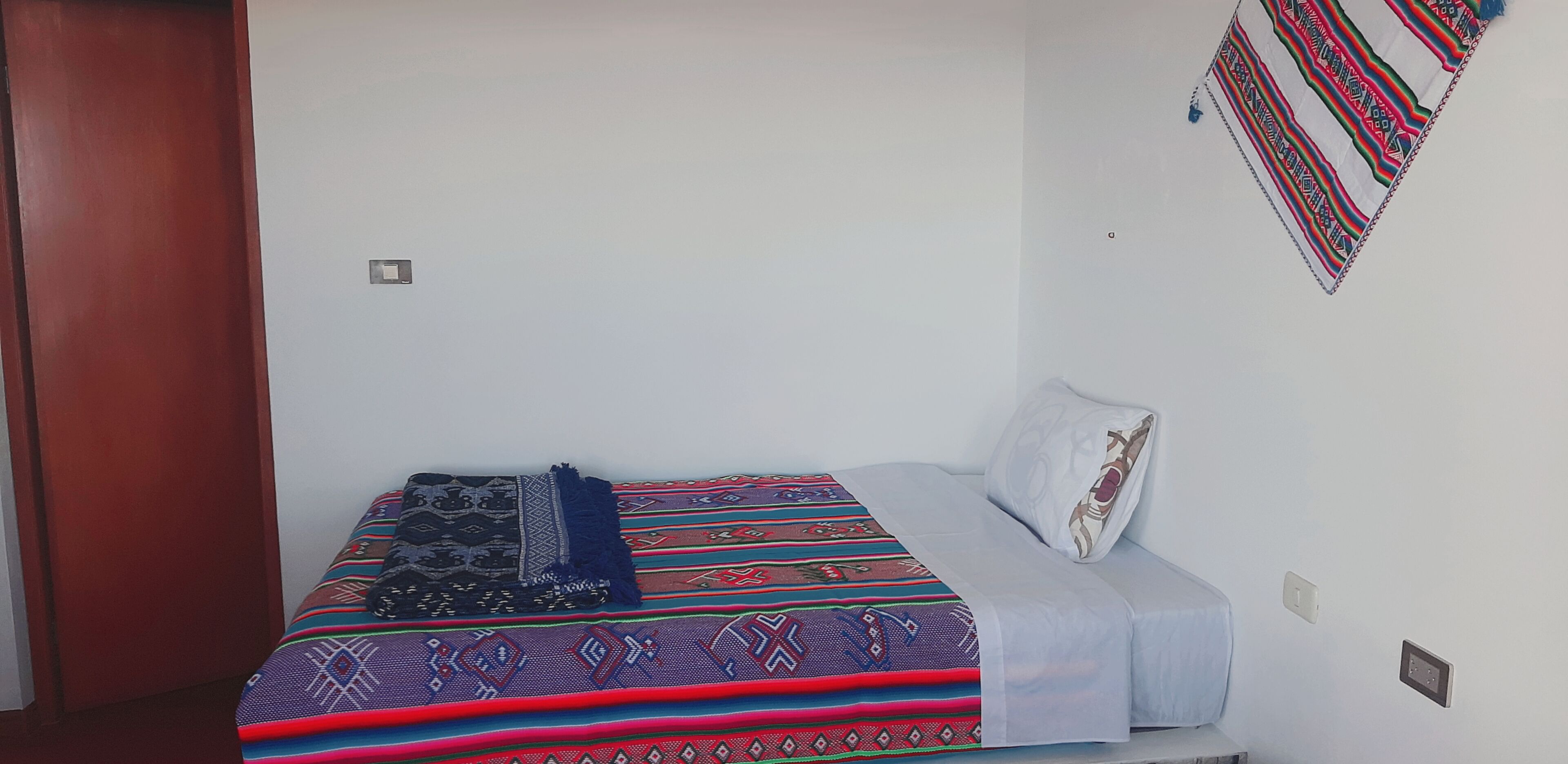 A single bed with a patterned bedspread and a pillow, a wall with a hanging textile, and a door.