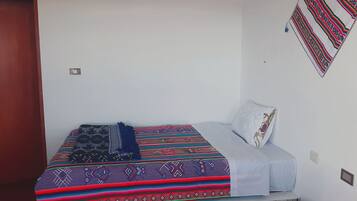 A single bed with a patterned bedspread and a pillow, a wall with a hanging textile, and a door.