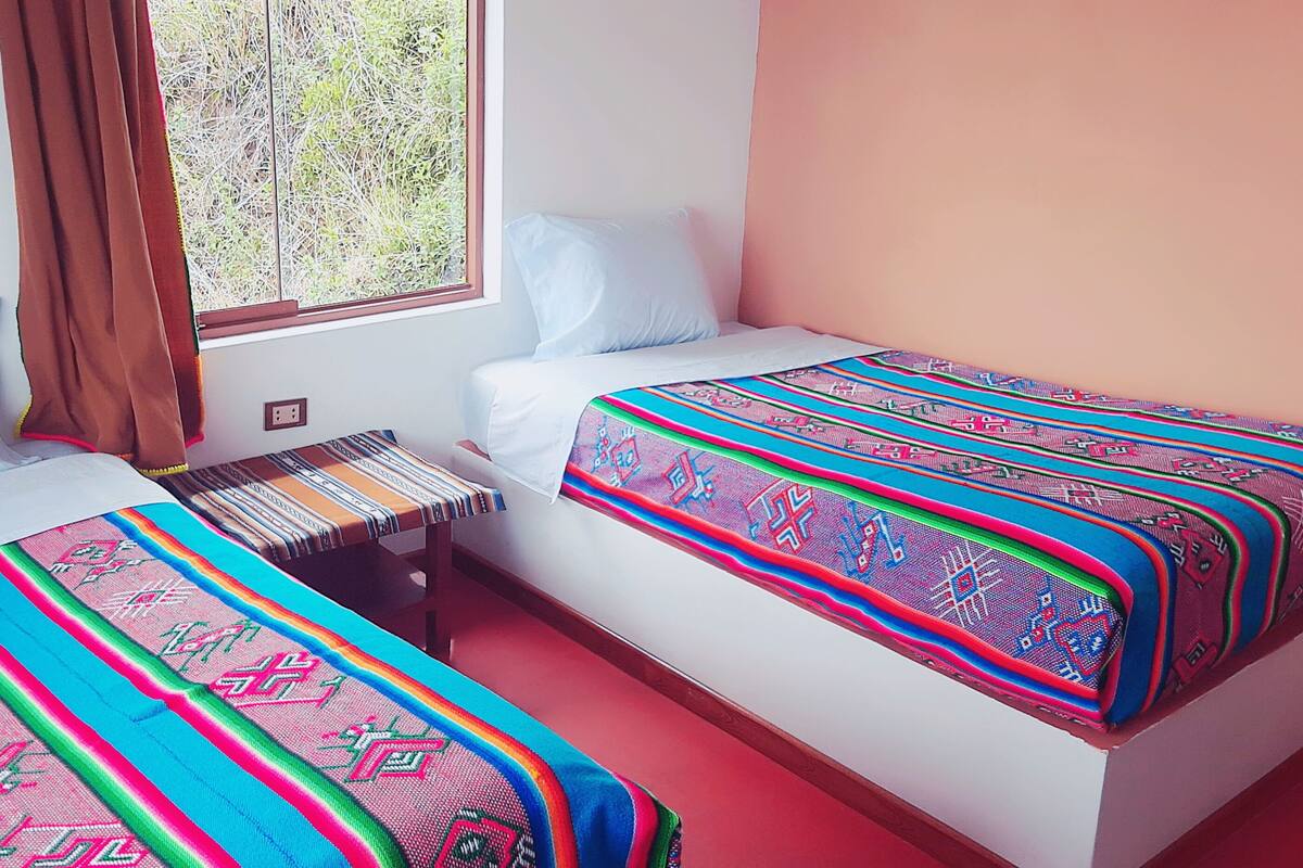 A room with two beds, each with a colorful blanket, a small table, and a window with curtains.