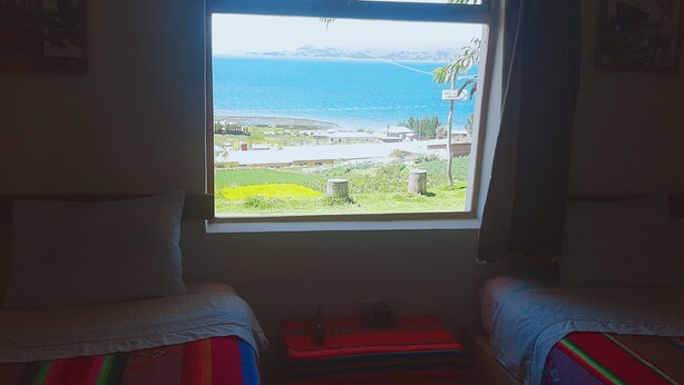 Une chambre avec deux lits, une fenêtre donnant sur la mer et une ville côtière, et une petite table recouverte d’une nappe rouge.