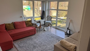 Un salon avec un canapé rouge, un fauteuil beige et une petite table ronde. Deux fenêtres offrent une vue sur l’extérieur.