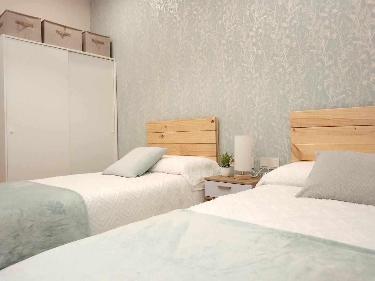 A bedroom with two beds, a wardrobe, and a nightstand with a lamp and a plant.