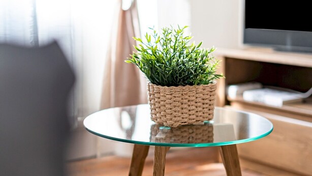 A round glass table with a woven plant pot on top, wooden legs, and a TV in the background. - The Loft - Beautiful flat in Chelsea, London - Pass the Keys (London)
