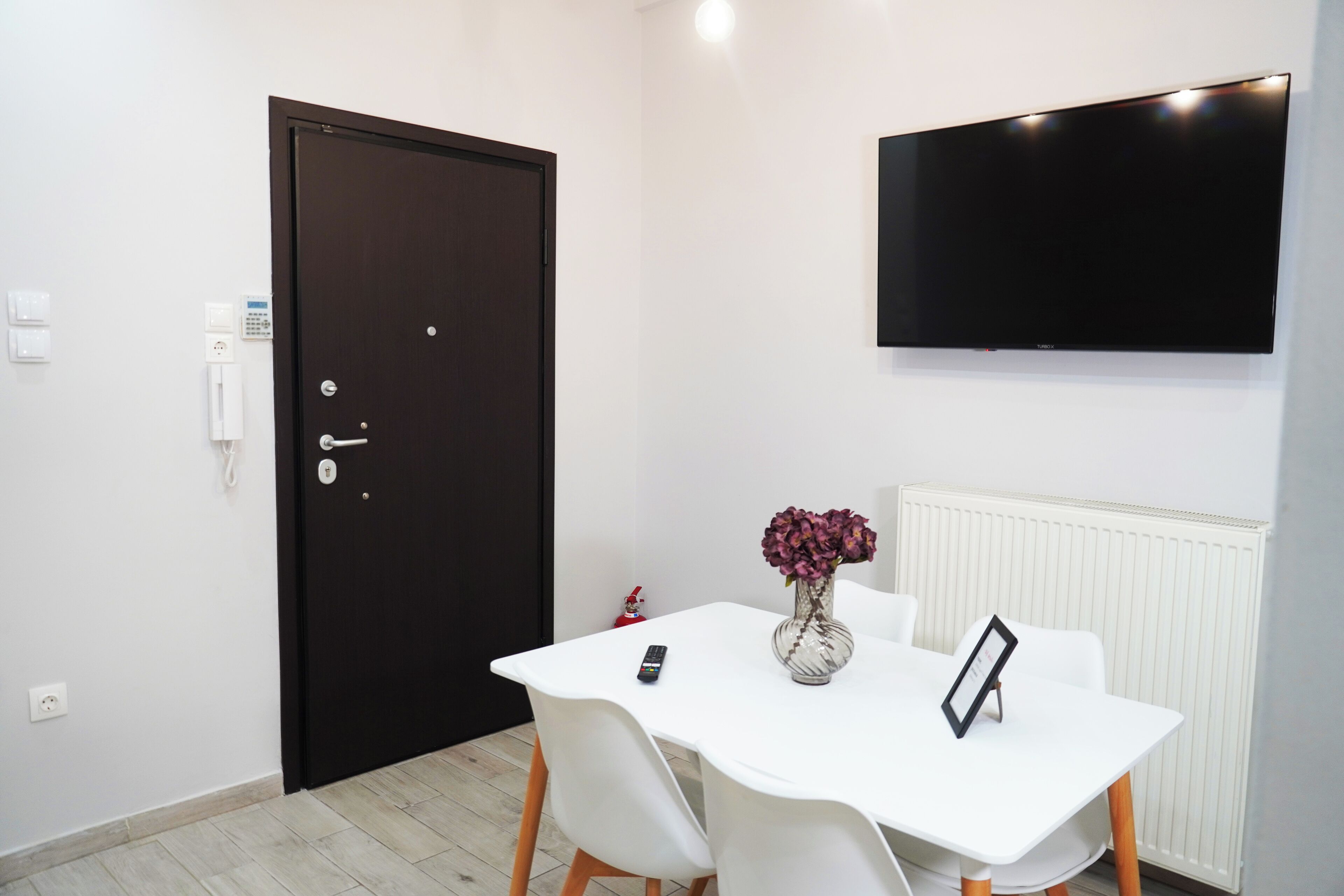 Ein Zimmer mit einem weißen Tisch, zwei weißen Stühlen, einer dunklen Tür, einem an der Wand befestigten Fernseher und einer Vase mit Blumen.
