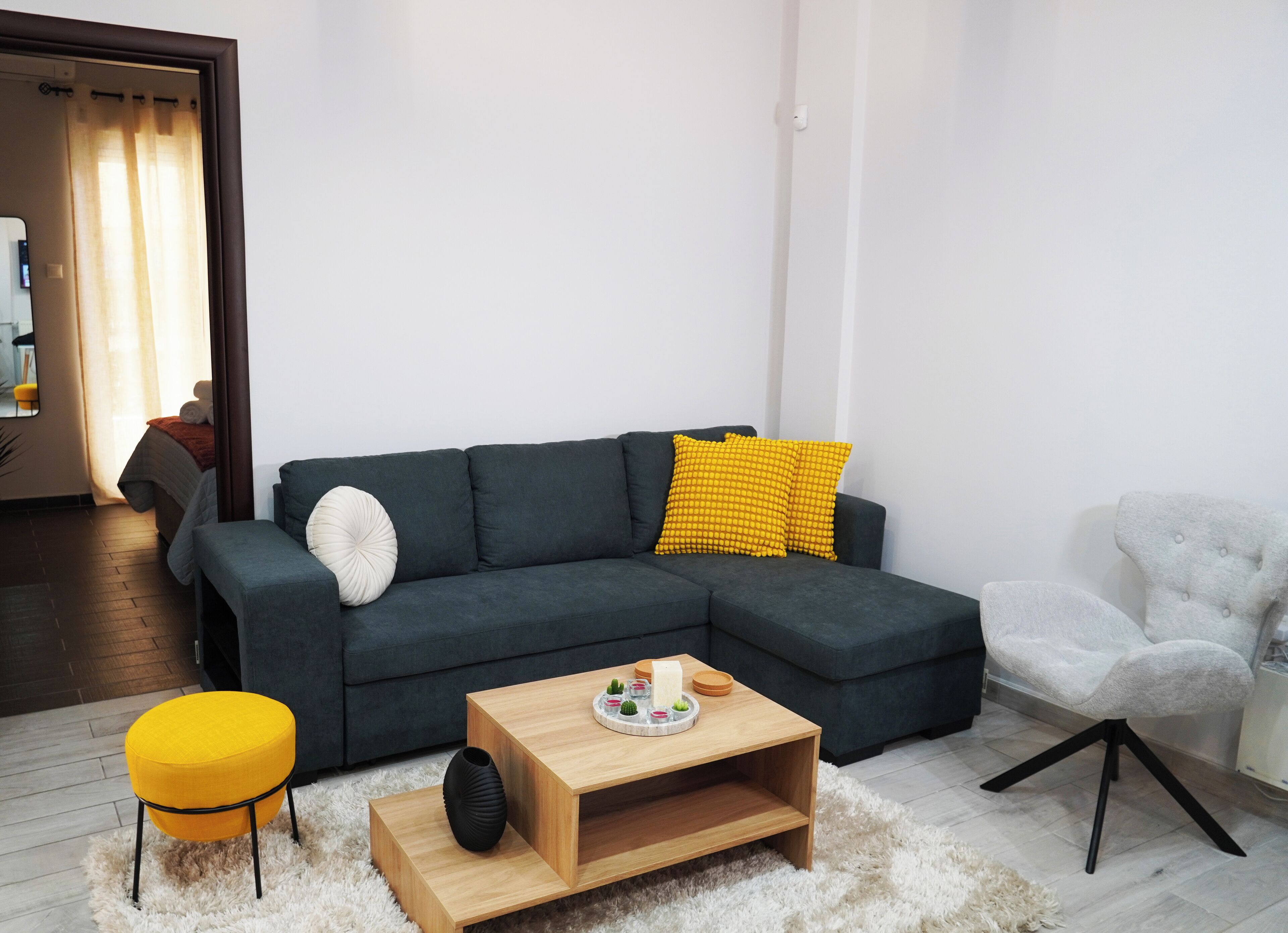 A modern living room with a dark grey sectional sofa, a wooden coffee table, a yellow ottoman, and a white armchair.