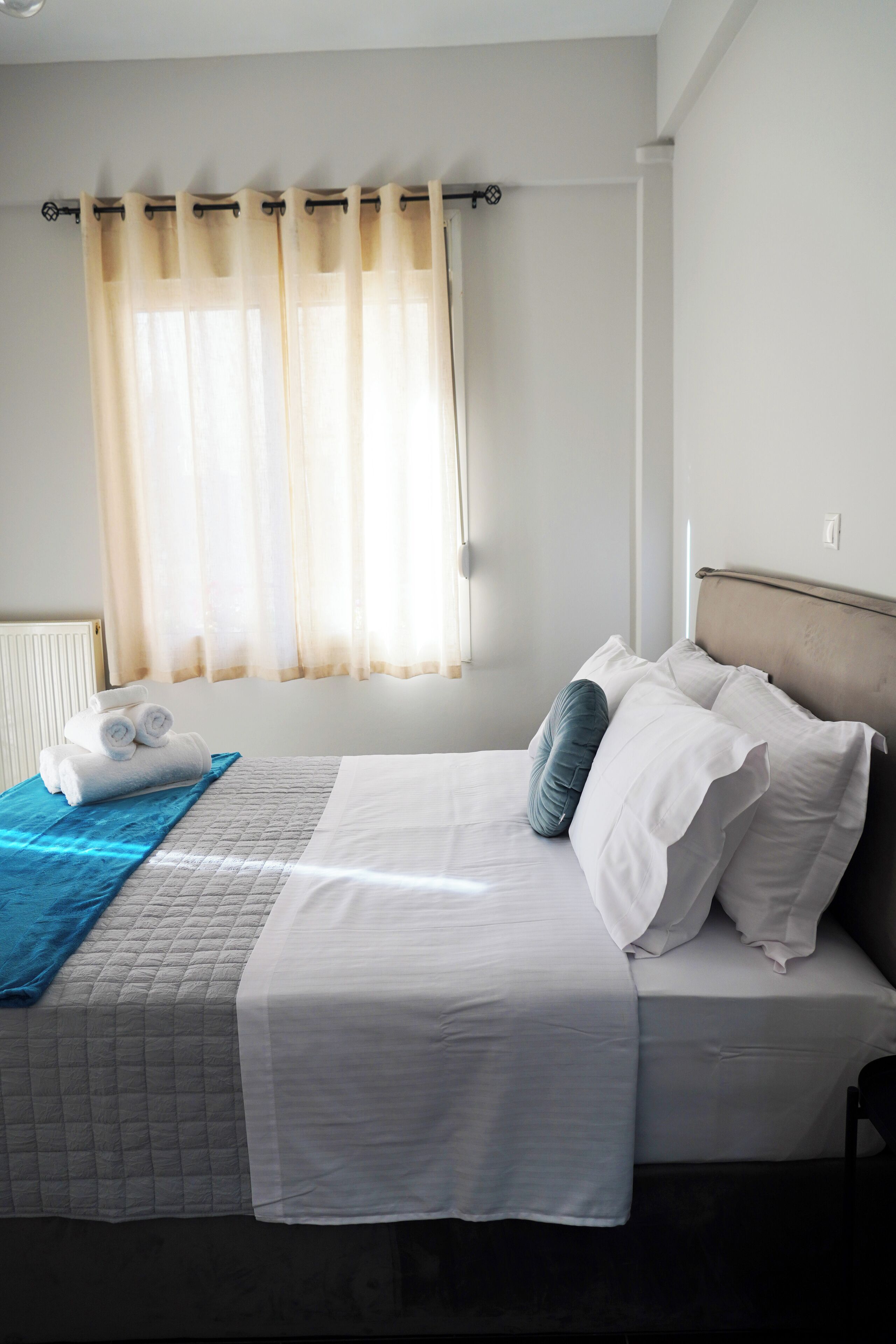 Un lit bien fait, avec des draps blancs et une couverture bleue, une tête de lit et une fen être ornée de rideaux légers.