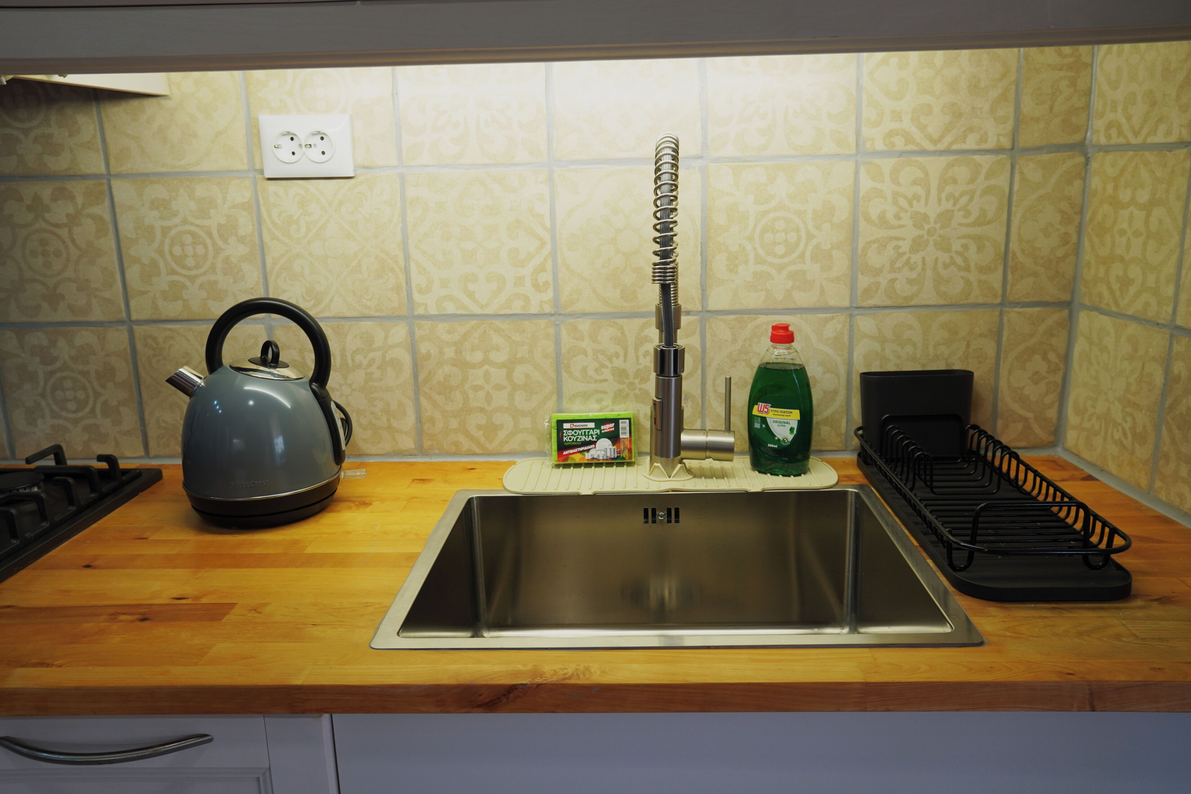 A kitchen with a stainless steel sink, a kettle, a dish rack, and a bottle of dish soap.