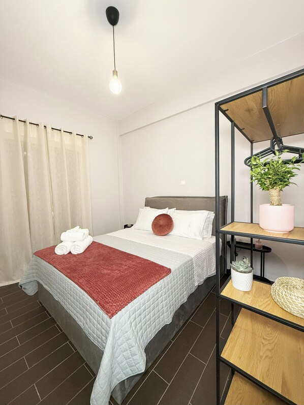A neatly made bed with a red blanket, a hanging plant, and a shelf with a plant and a candle. - Athenian Petralona Suite (Athens)