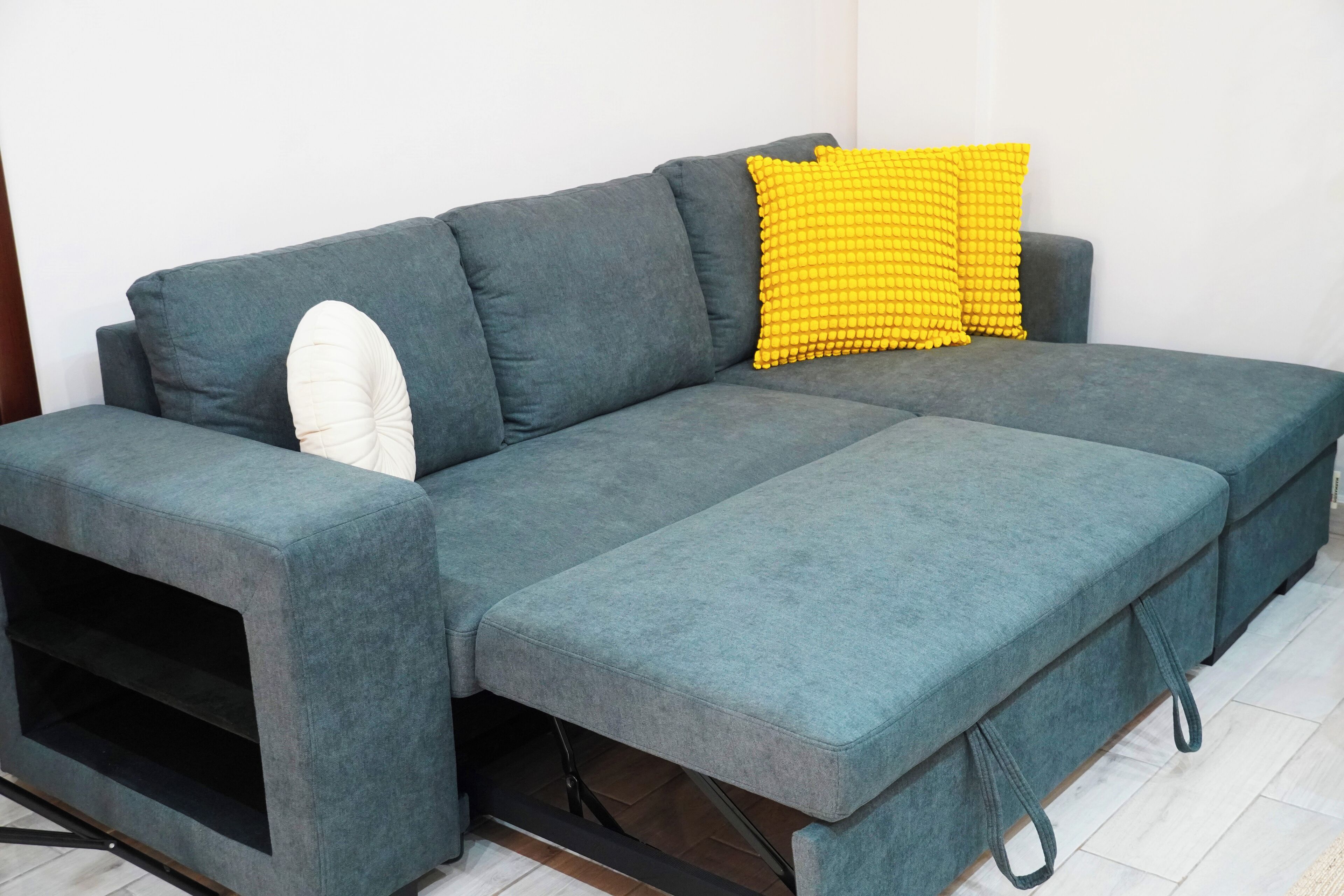 A grey sectional sofa with a white and yellow pillow, a black shelf, and a grey bench.
