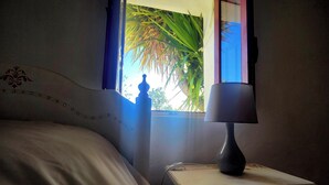 Una cama con cabecera, una mesita de noche con lámpara y una ventana con vista a follaje.