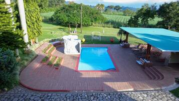 A swimming pool with a red border, surrounded by a wooden deck with a stone pathway leading to it. There is a covered seating area with a blue roof and a view of a lush green landscape in the background.