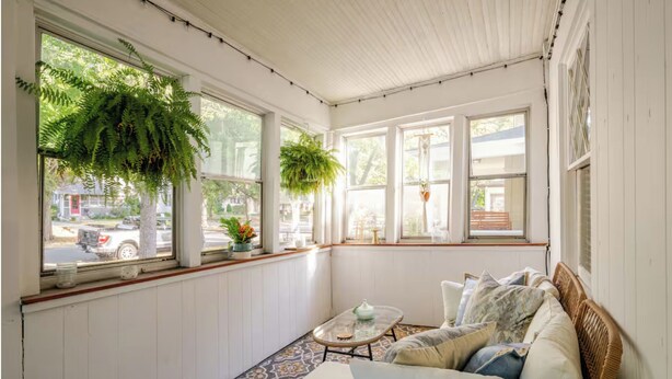 A sunroom with large windows, a wicker sofa, a patterned floor, and hanging plants.
