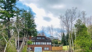A two-story house with a dock extending into a lake, surrounded by trees and a clear sky.