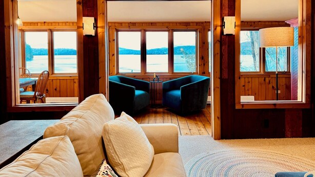 A cozy cabin interior with a sofa, two armchairs, and a dining table. Large windows offer views of a snowy landscape.