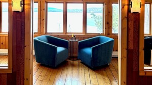 A room with wooden walls, two blue armchairs, a small table, and large windows offering a view of the outdoors.