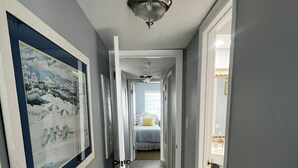 A hallway with a framed picture on the left, a bed with a yellow pillow in the distance, and a bathroom visible through an open door. - Driftwood Towers 2 bedroom unit 2C. Gulf front condo in Gulf Shores Alabama. (Gulf Shores)