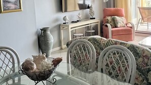 A living room with a ceiling fan, a TV, a pink armchair, a dining table with chairs, and a vase on the table. - Driftwood Towers 2 bedroom unit 2C. Gulf front condo in Gulf Shores Alabama. (Gulf Shores)
