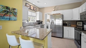 A kitchen with white cabinets, a granite countertop, a stainless steel refrigerator, and a painting of a palm tree. - Quiet Retreat, Golf Cart Included, Pet Friendly - 603 Porpoise (Fripp Island)