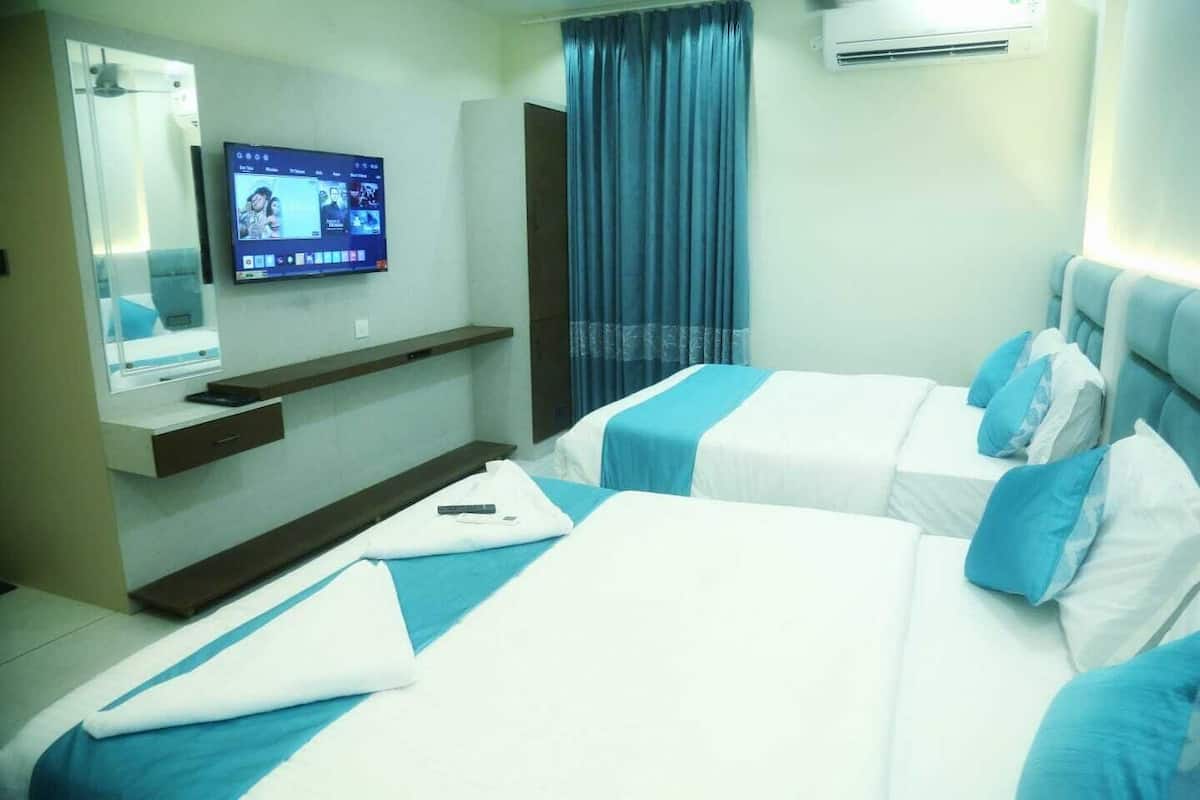 A hotel room with two beds, a TV mounted on the wall, a small desk with a chair, and a window with curtains.