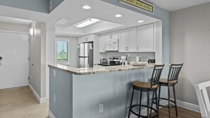 A modern kitchen with white cabinets, a marble countertop, and a built-in microwave. There are two bar stools and a window with blinds. - Perfect Beach Vacation -Sanibel Island's Loggerhead Cay 234 (Sanibel)