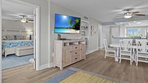 A spacious living room with a large flat-screen TV, a dining area with white furniture, and a kitchen with a ceiling fan. - Perfect Beach Vacation -Sanibel Island's Loggerhead Cay 234 (Sanibel)