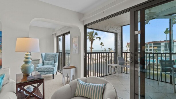 A modern living room with a sofa, armchair, and a small table with a lamp. There is a balcony with a glass railing and a view of the ocean and palm trees.