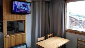 A room with a wooden table and chairs, a TV mounted on the wall, a window with curtains, and a view of buildings outside.