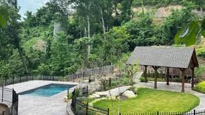 Une piscine entourée d’une clôture, d’un belvédère et d’une pelouse avec des arbres en arrière-plan.