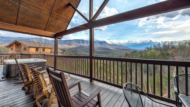 Une terrasse en bois avec un jacuzzi, des chaises en bois et une table offrant une vue sur un paysage montagneux.