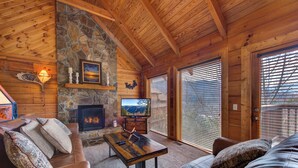 L’intérieur d’un chalet confortable, avec des murs en rondins de bois, une cheminée en pierre, des meubles en cuir et une grande fenêtre équipée de stores.