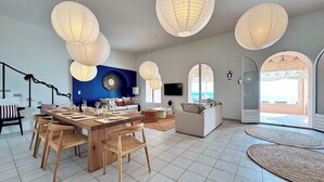 Amplia sala de estar con mesa de comedor preparada para seis comensales, un sofá y un televisor montado en la pared. Amplias ventanas ofrecen vistas al exterior.