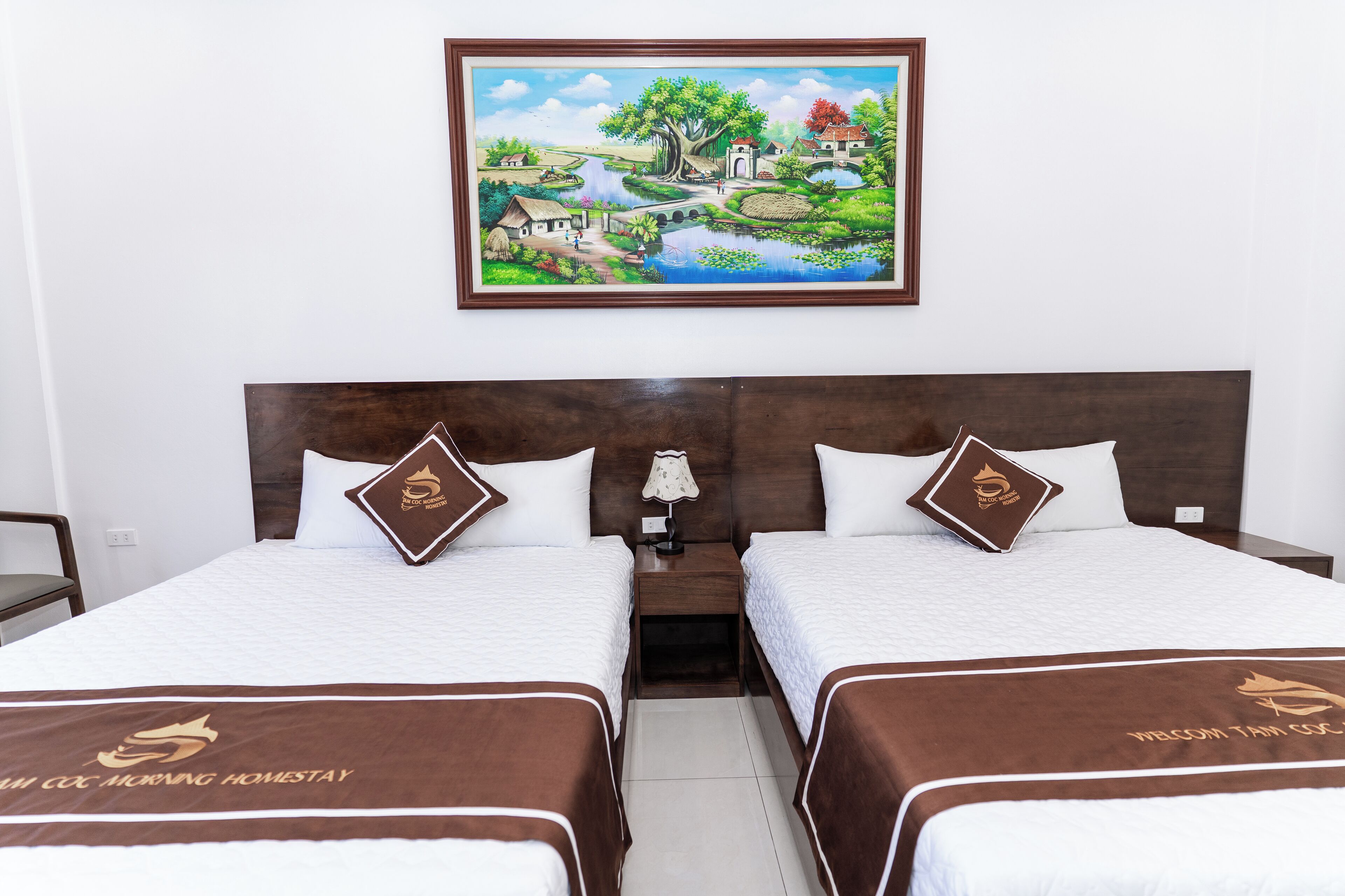 A hotel room with two beds, a nightstand, a lamp, and a painting of a village scene on the wall.