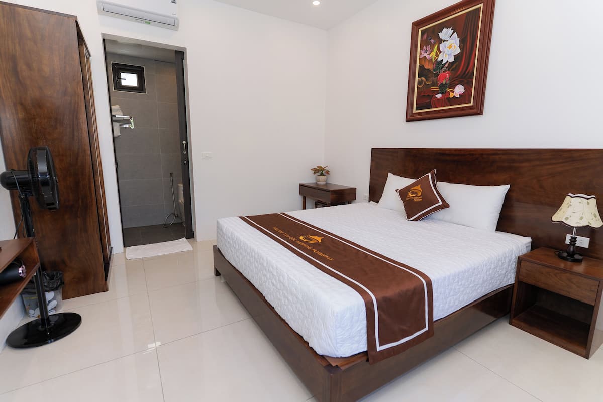 A neatly made bed with a brown headboard and a white comforter, a small bedside table with a lamp, and a framed floral artwork on the wall.
