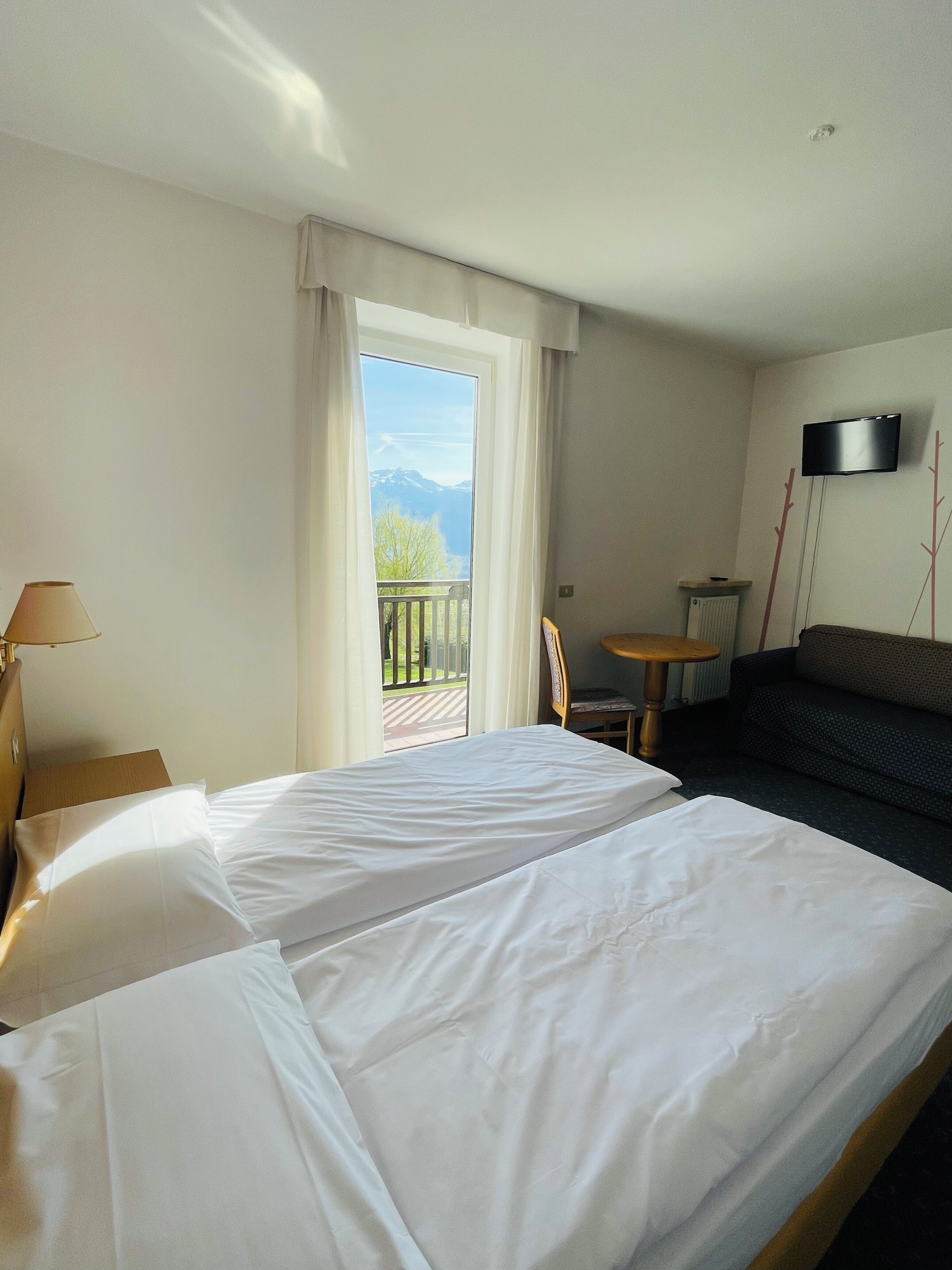 A hotel room with a large bed, a small table, a sofa, a TV, and a balcony with a view of mountains.