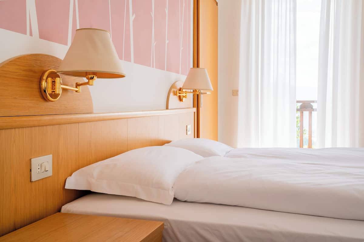 A hotel room with a wooden headboard, two bedside lamps, a bed with white linens, and a window with sheer curtains.