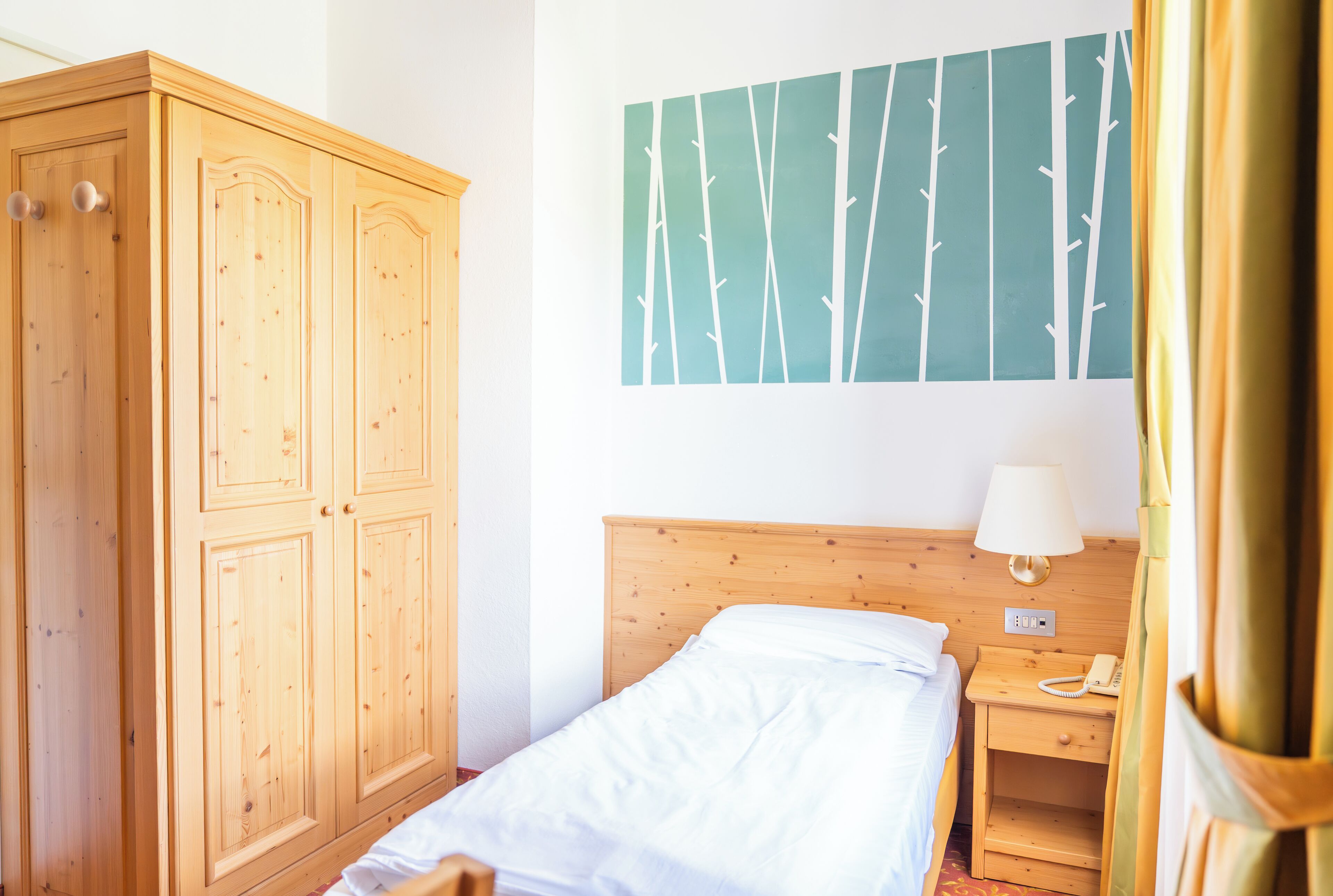 Ein Hotelzimmer mit Holzschrank, einem Bett mit weißer Bettwäsche, einem Nachttisch mit Lampe und einem Baum-Wandbild.