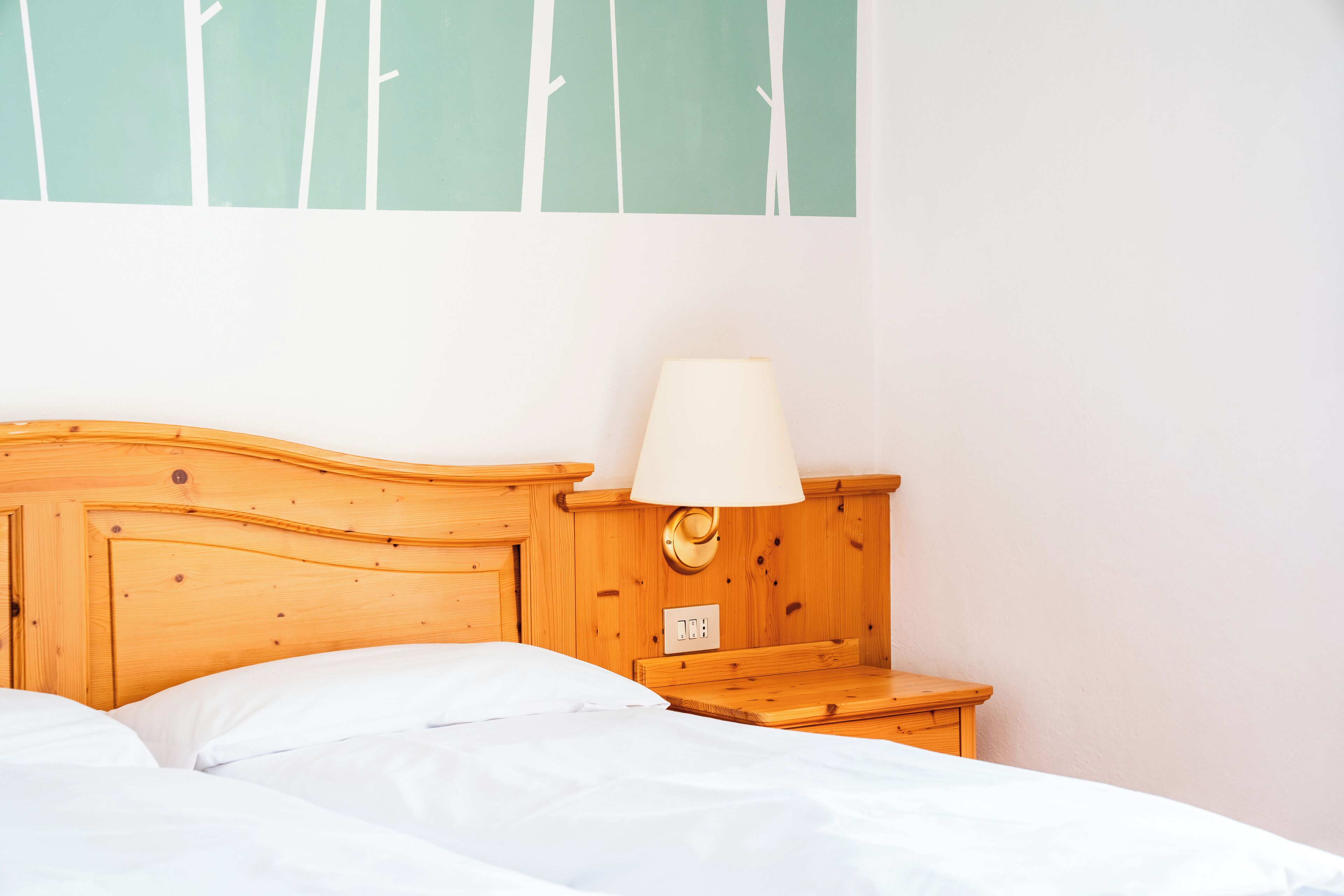 Ein ordentlich bezogenes Bett mit weißen Laken, ein Nachttisch aus Holz mit einer Lampe und eine Wand mit einem schlichten Grün-Weiß-Muster.