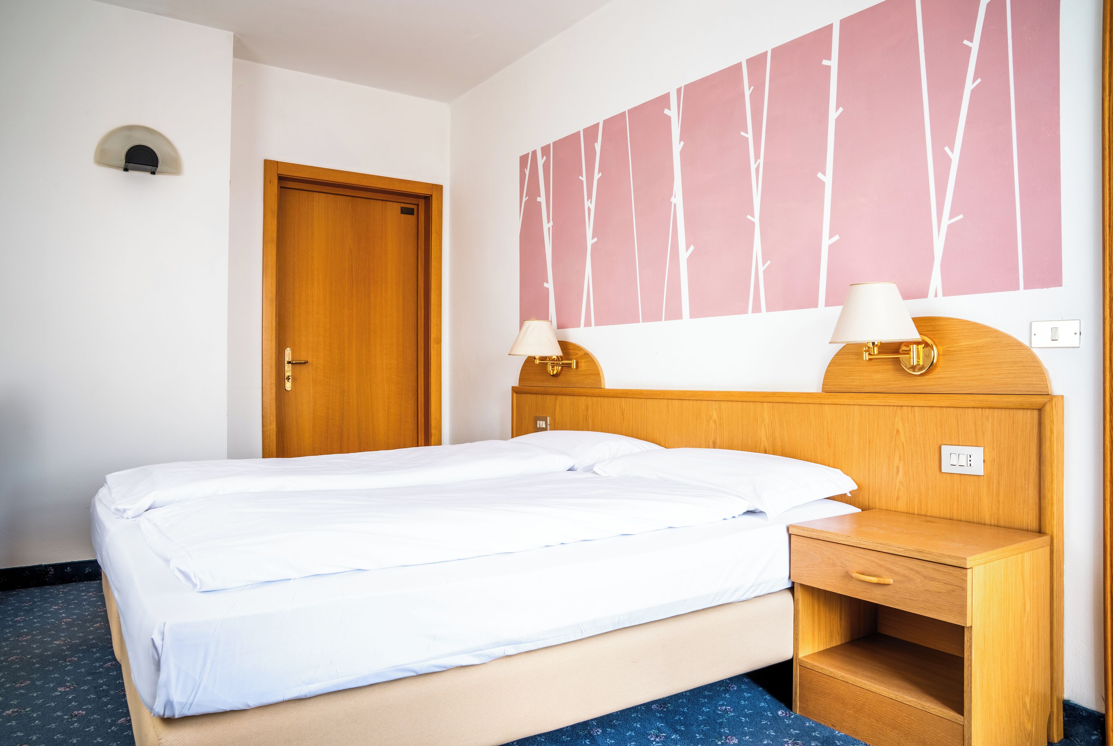Ein Hotelzimmer mit einem Bett, einem Holzkopfteil, Nachttischlampen, einem Nachttisch und einer Wand mit Baummuster.