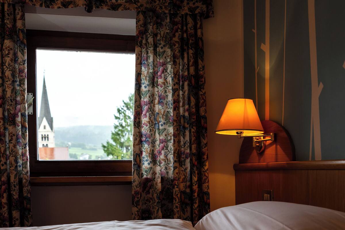 A hotel room with a bed, a window with floral curtains, and a lamp on a bedside table.