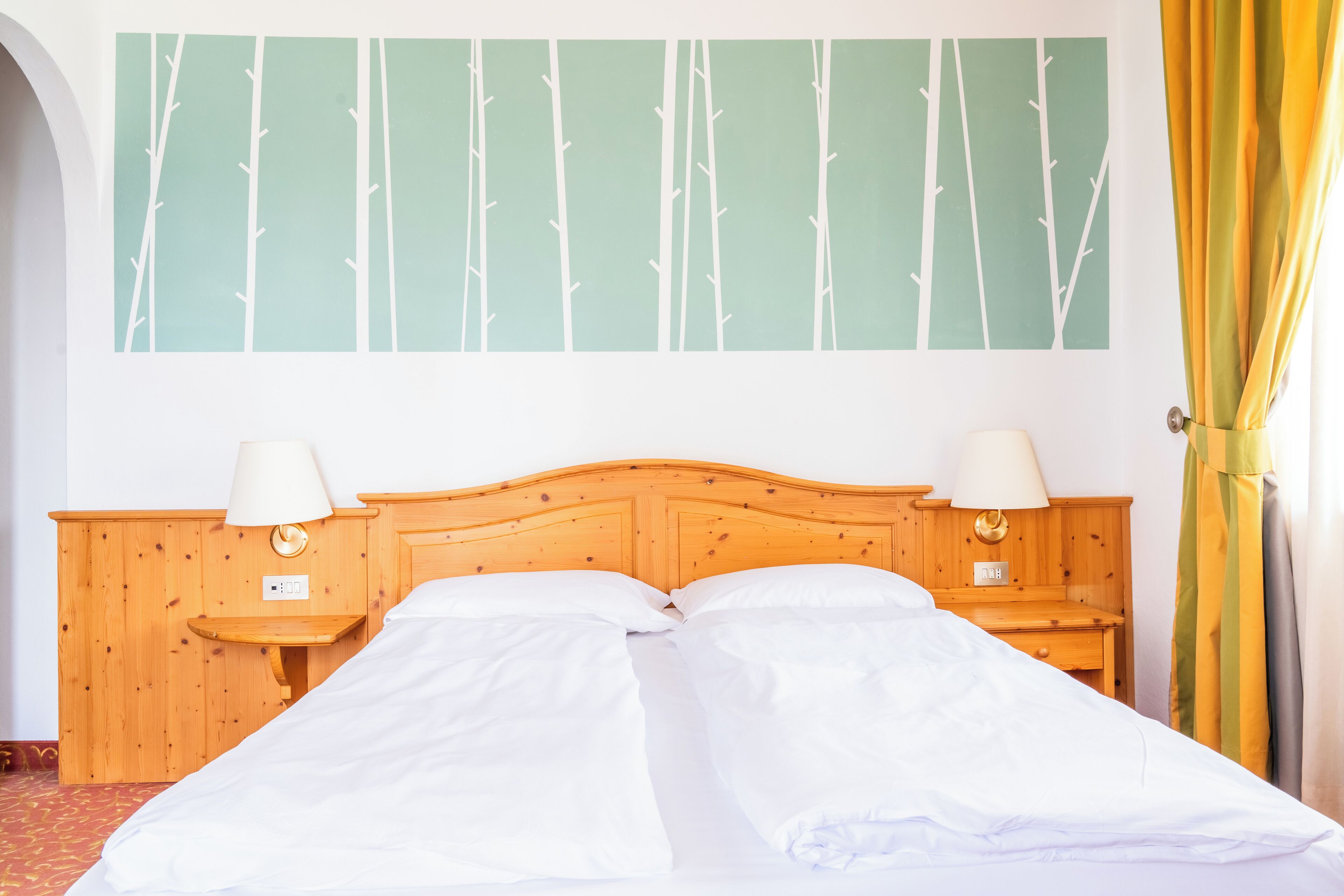 A double bed with white linens, two bedside tables with lamps, a wooden headboard, and a wall with a bamboo forest mural.