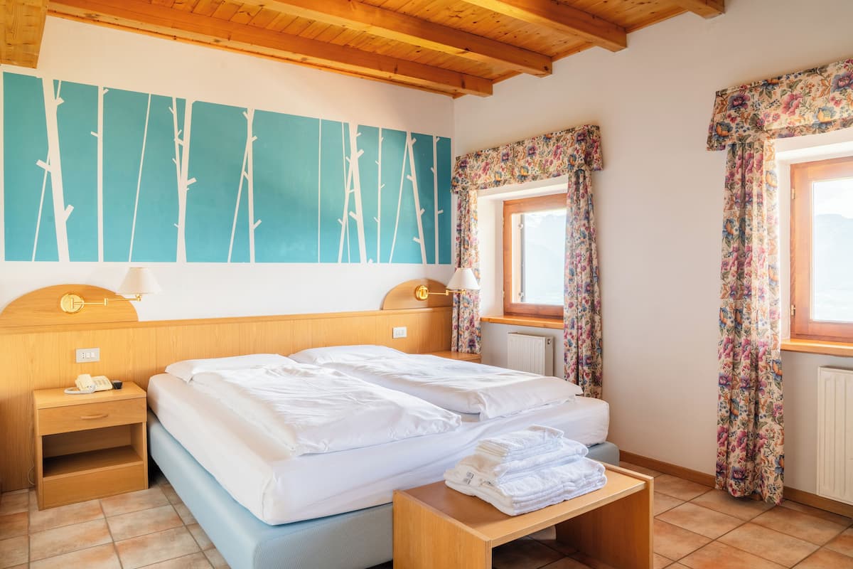 A bedroom with two beds, a wooden headboard, a nightstand with a lamp, a window with floral curtains, and a wall with a tree design.